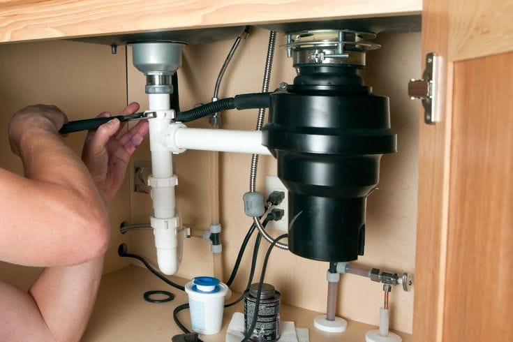Plumber installing a garbage disposal beneath a kitchen sink in Seaside